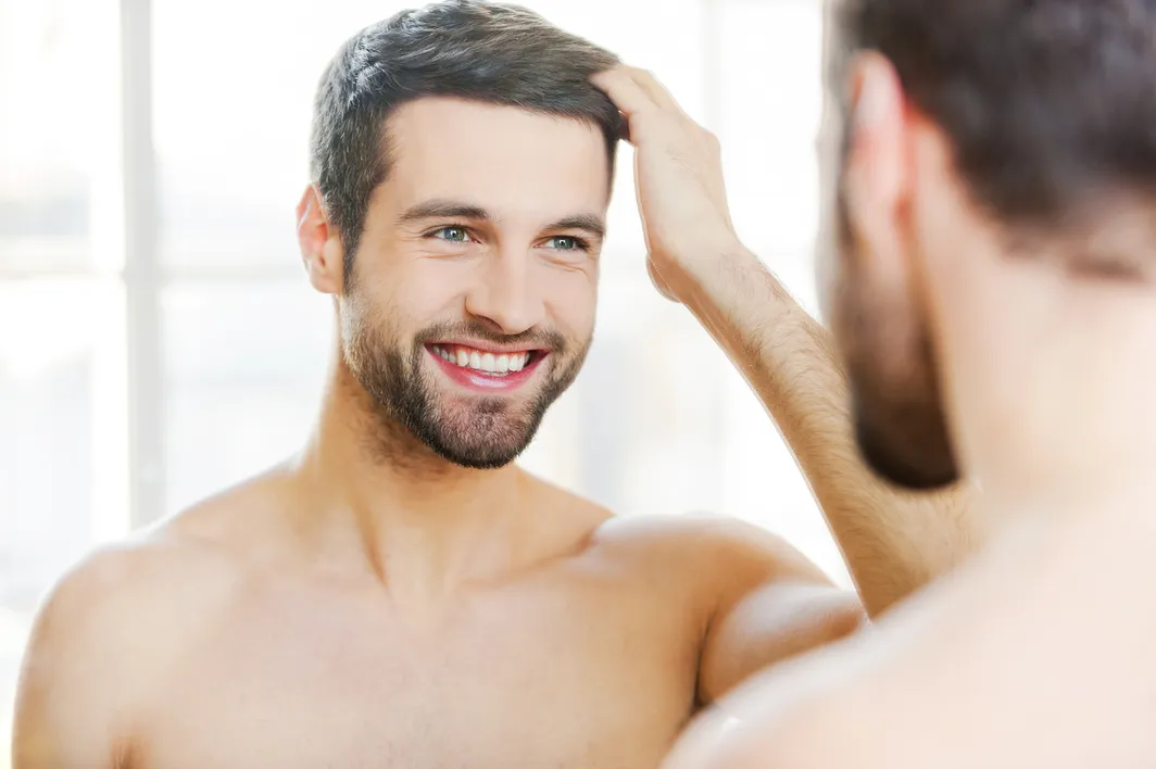 Smiling man checking his dense hairline in the mirror, evaluating a minoxidil vs hair transplant treatment.