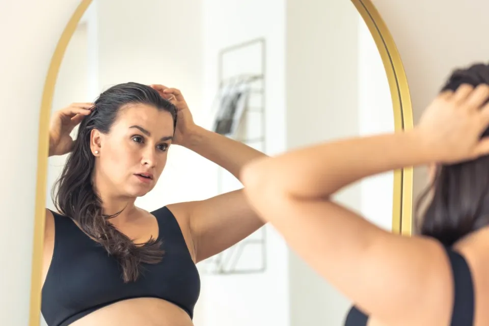 Woman looking at her front hairline in the mirror, experiencing the first visible signs of postpartum hair loss.