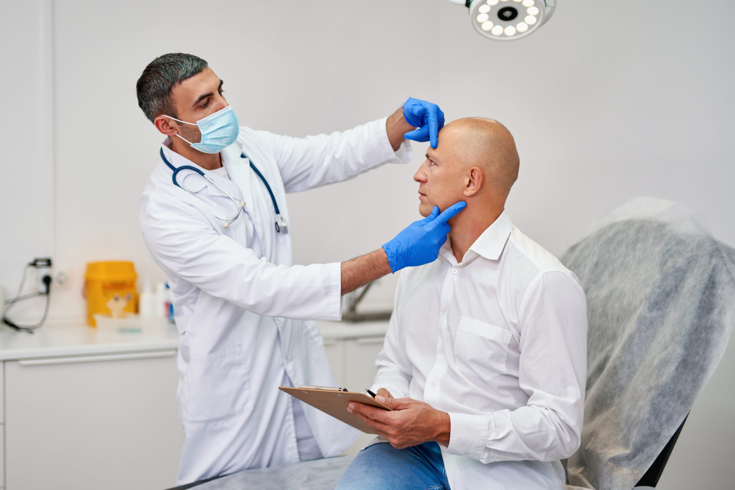A doctor in a mask examines a man's bald scalp during a consultation to discuss "does hair transplant hurt".