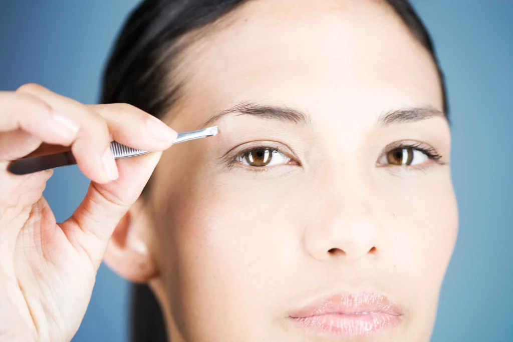 Woman using tweezers on her brow, highlighting the need or preparation for an eyebrow hair transplant.