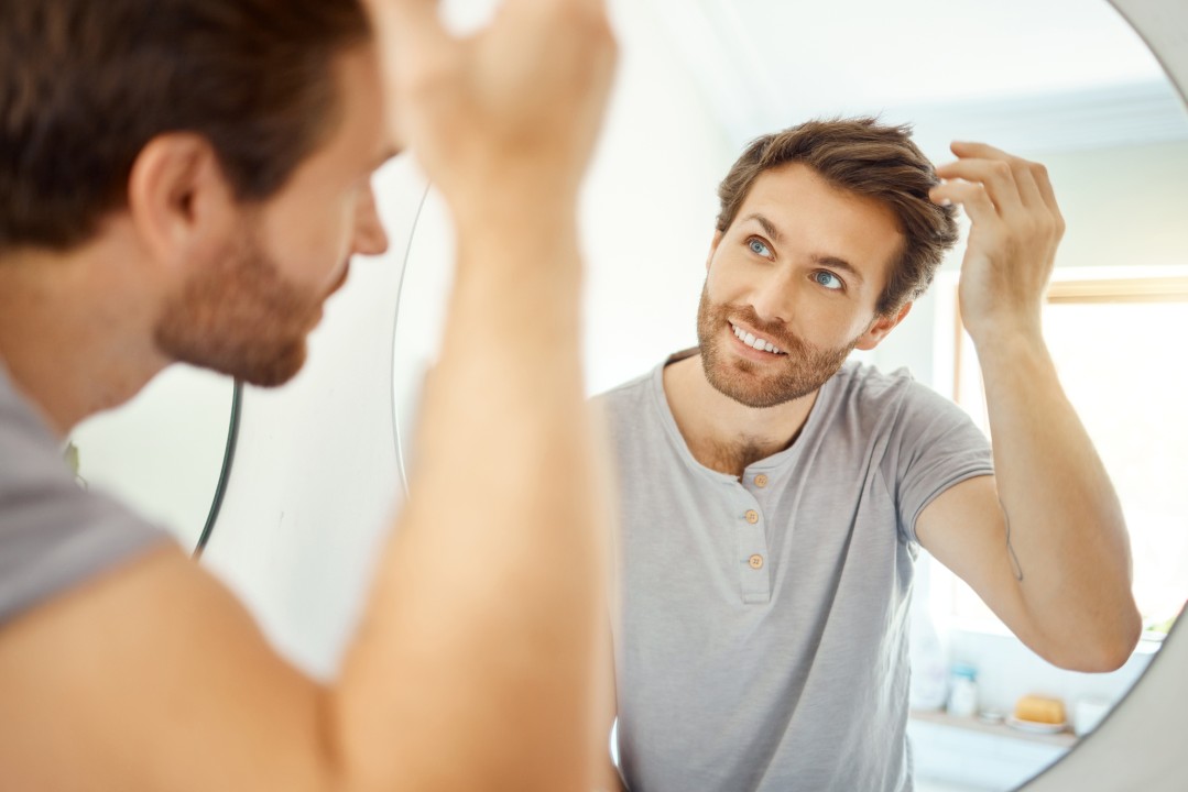Happy man checking his fuller hair in a mirror, a possible result of hair growth peptides treatment.